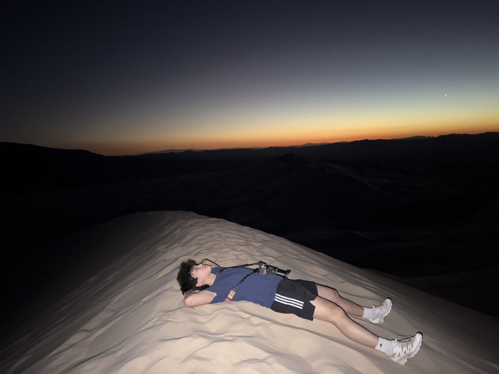 Winston lying on his back on top of a sand dune at dusk, desert ridges behind him.