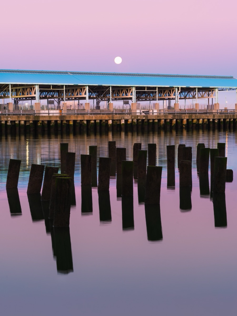 Full moon rising over a waterfront pier at dusk, weathered pilings reflected in still pink water