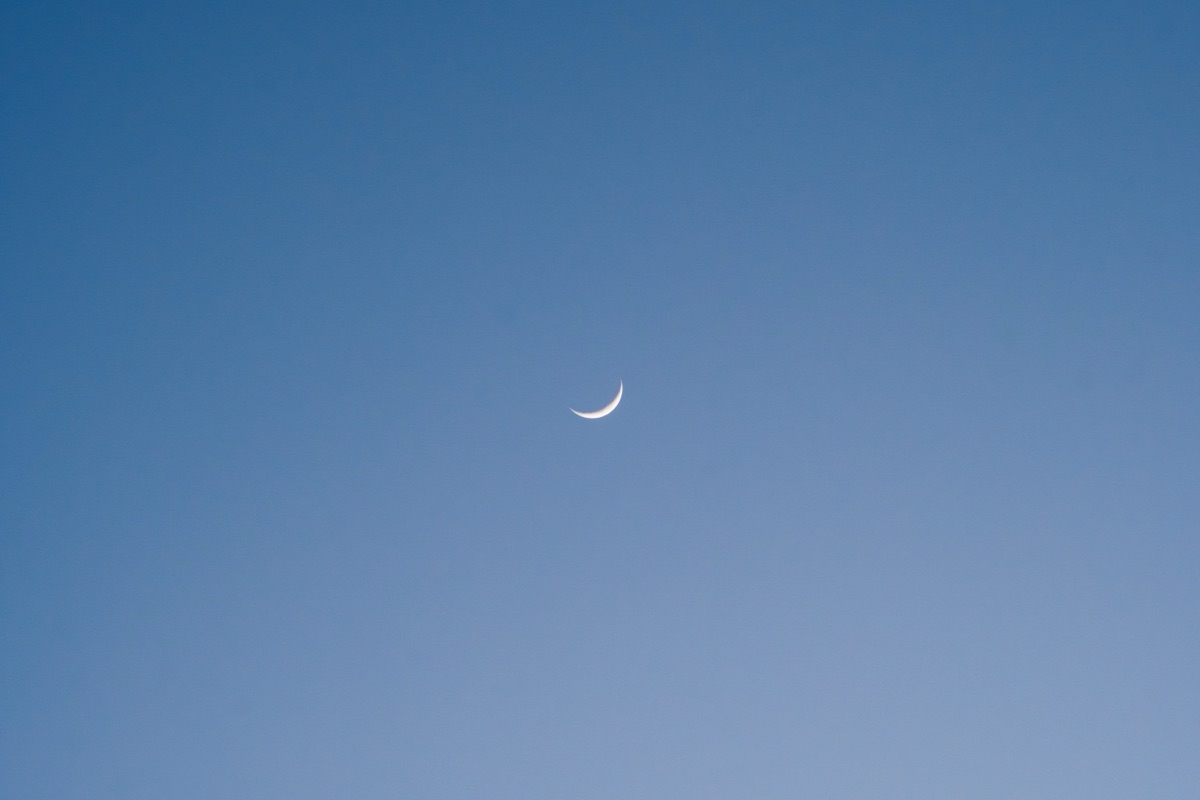 A slender crescent moon in a clear blue daytime sky