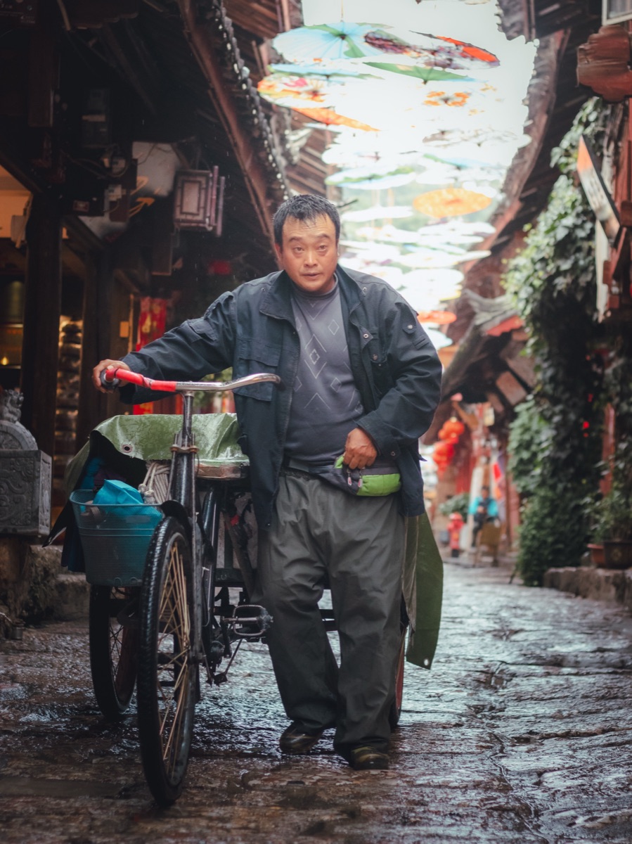 Man pushing his bicycle down a rain-slicked cobblestone lane beneath a canopy of painted umbrellas, Lijiang, China