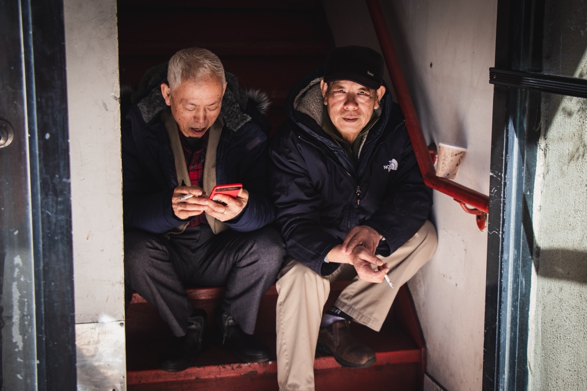 Two older men sharing a cigarette and a phone screen in a stairwell doorway on a cold day