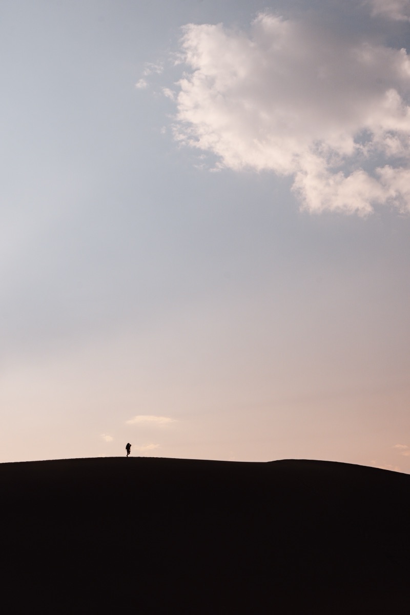 A solitary hiker cresting a dark dune ridge under a wide pastel sky at dusk
