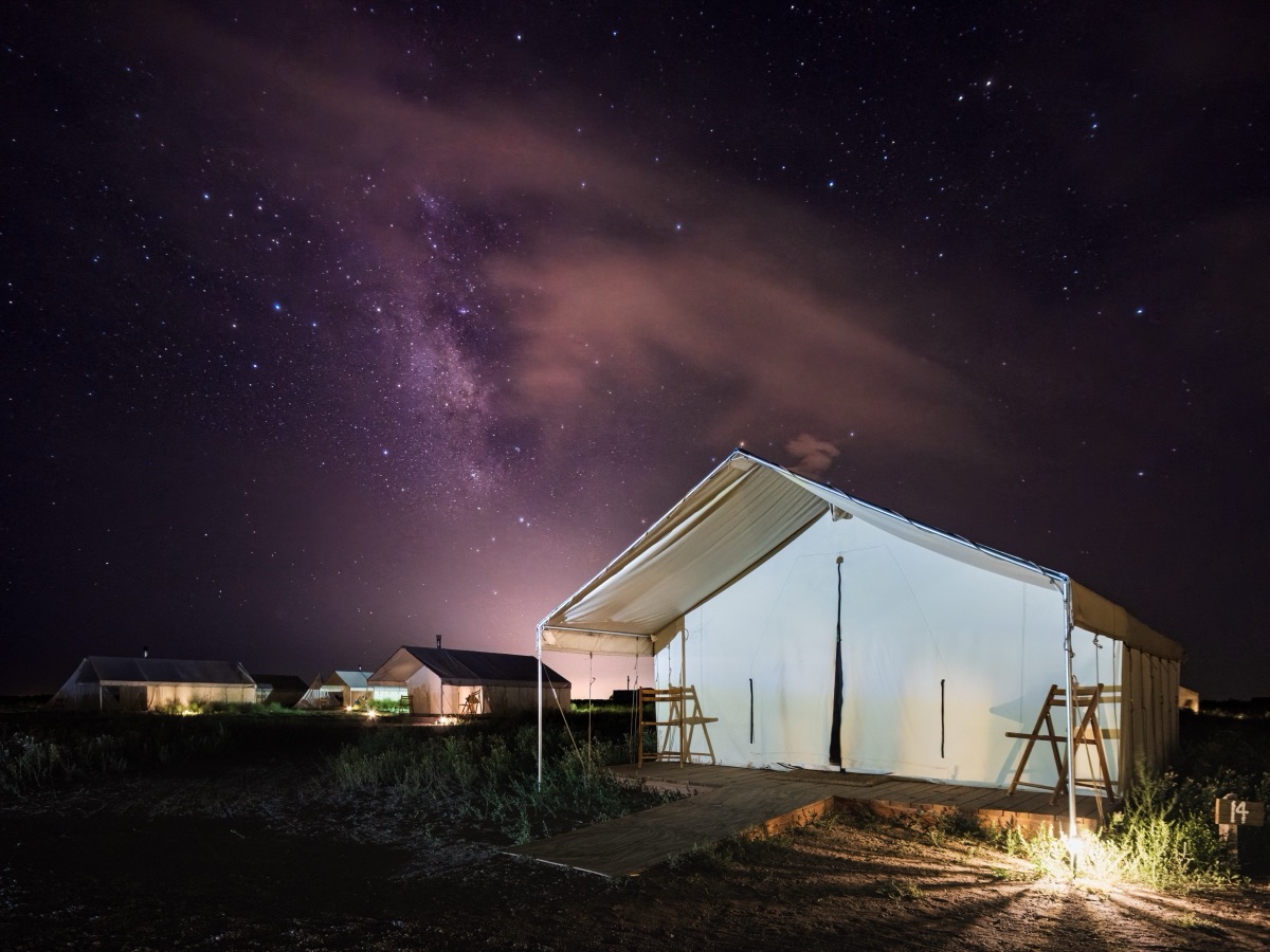 The Milky Way arching over a lit canvas tent at a remote desert camp
