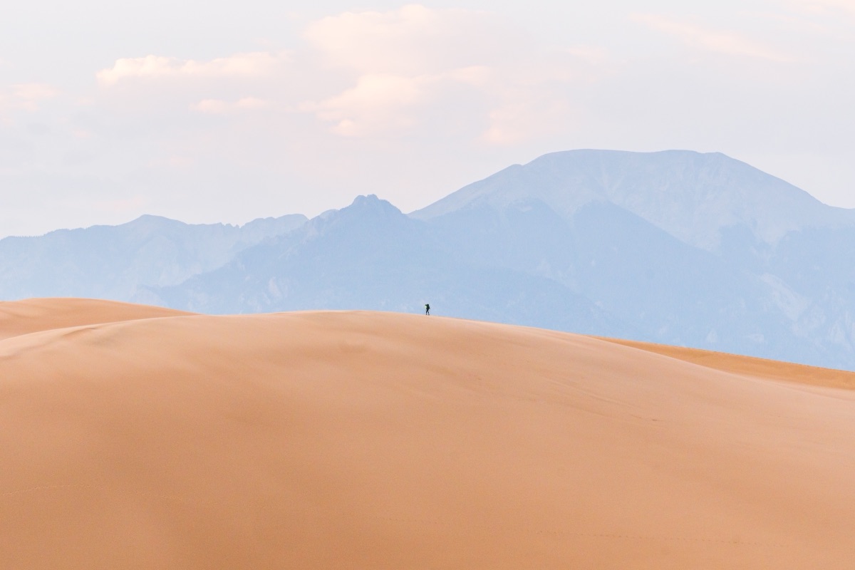 A lone figure walking the crest of a sand dune below hazy blue mountains, Great Sand Dunes, Colorado