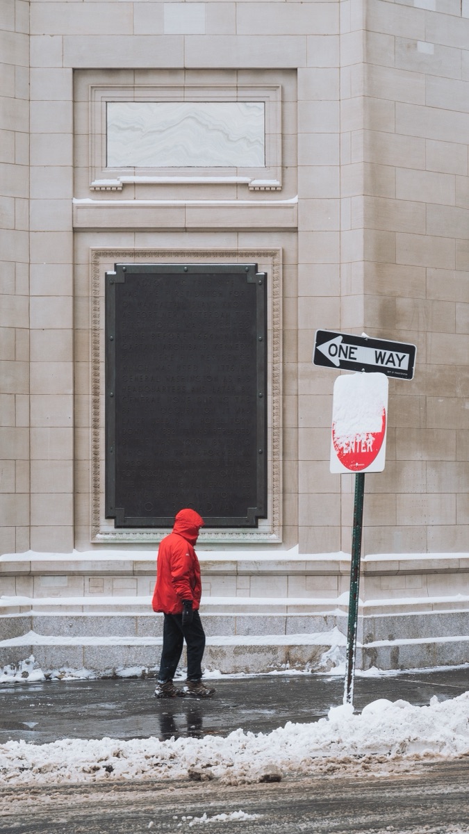 A figure in a red parka passing a limestone facade during a snowstorm