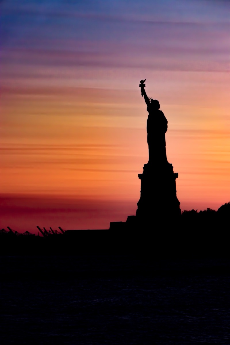 The Statue of Liberty silhouetted against a banded orange and violet sunset, New York Harbor