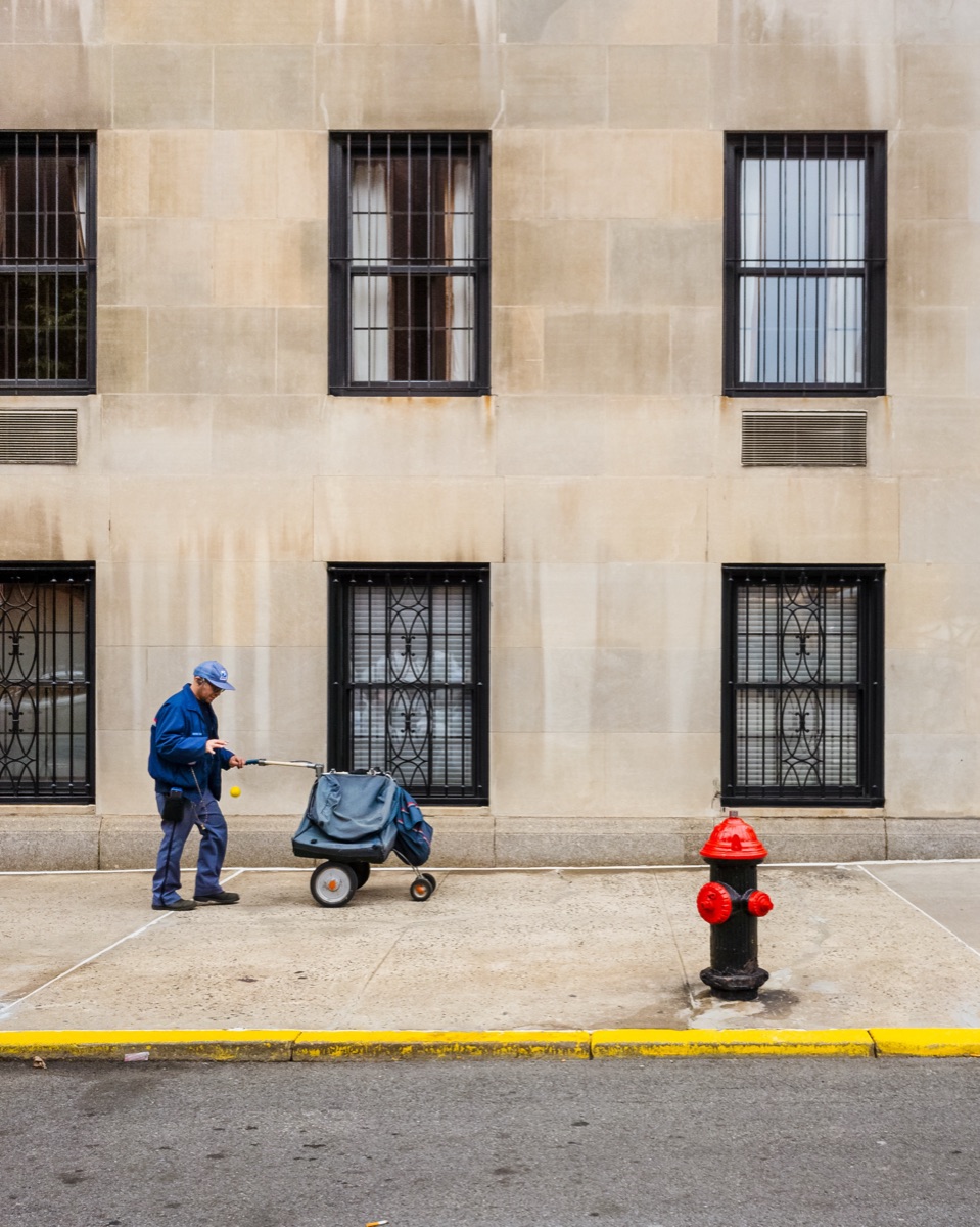 A sanitation worker pushing a cart past the barred windows of a stone building, New York