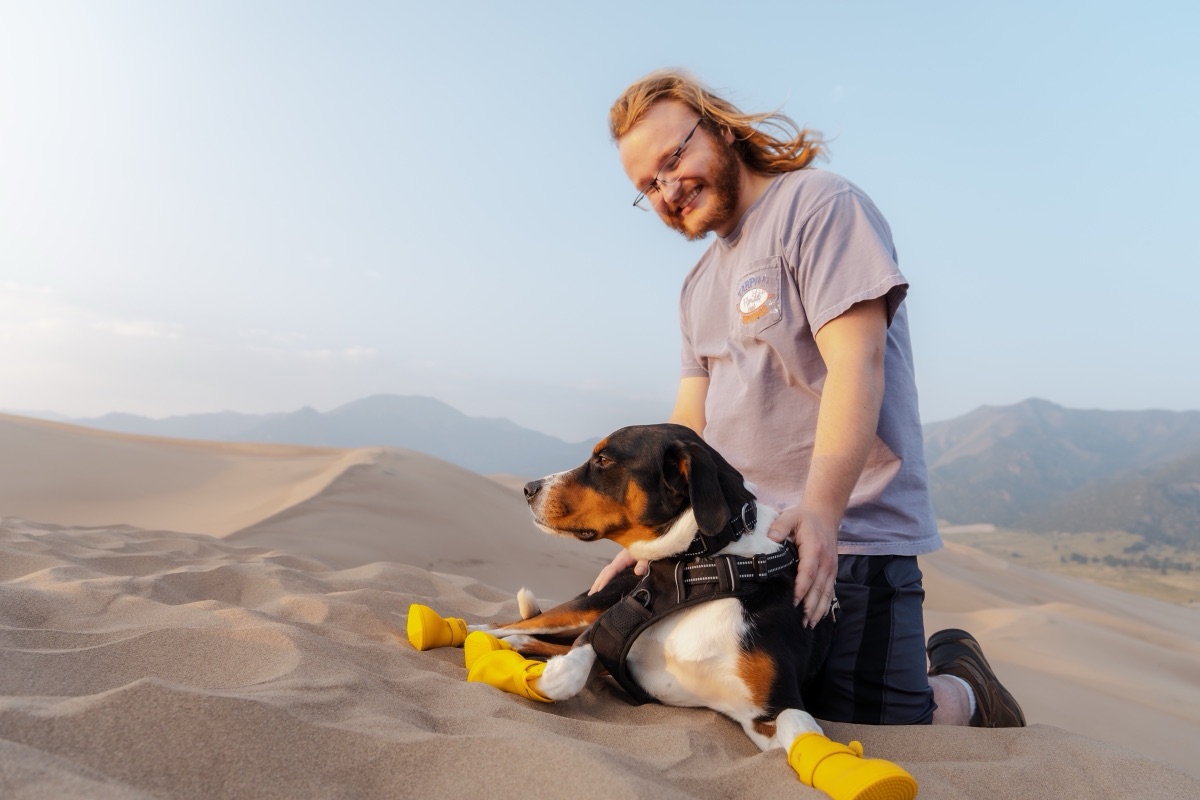 A hiker kneeling beside his dog in booties on the sand dunes at golden hour
