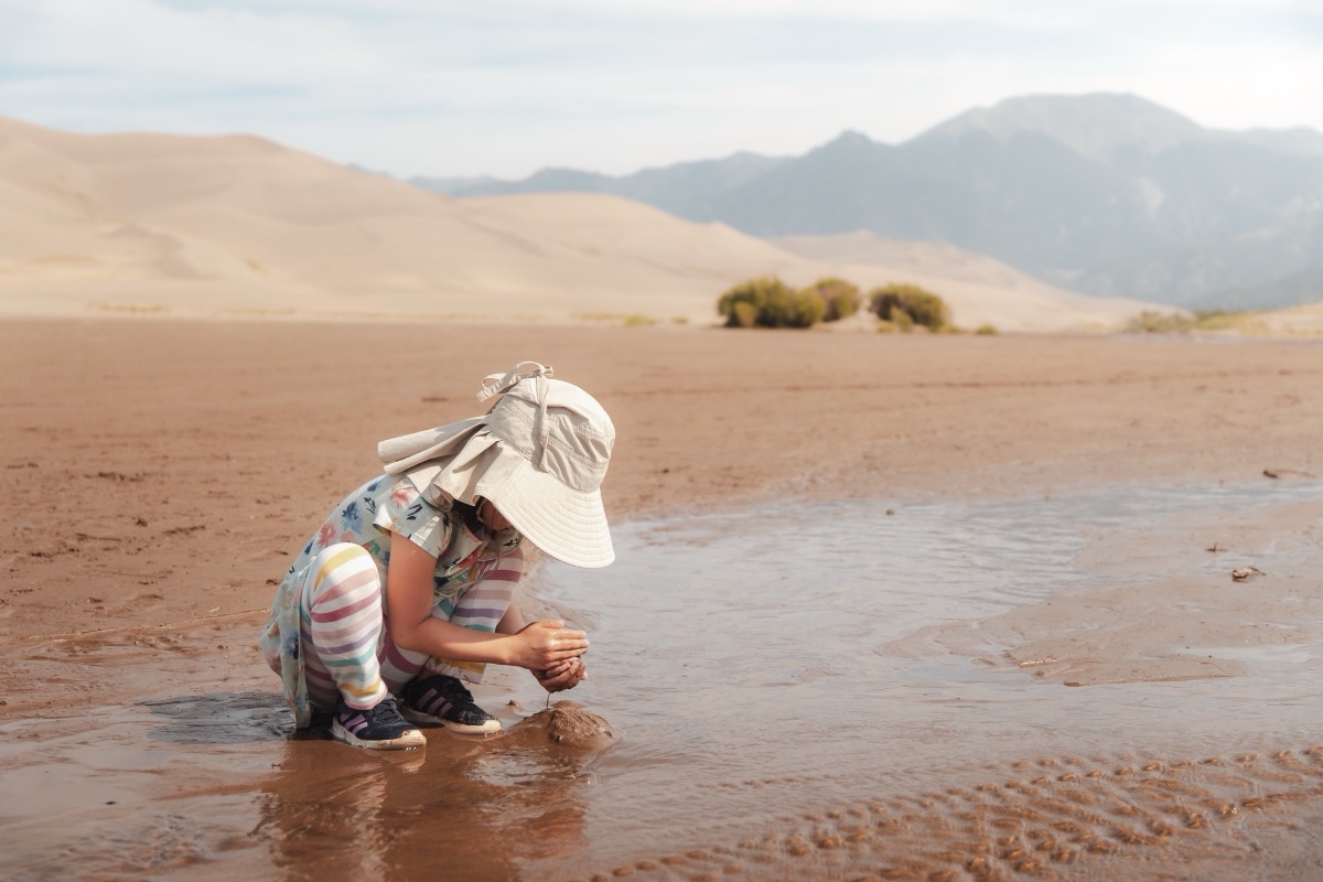 A small child in a sun hat crouching to play in a shallow creek below the dunes, Great Sand Dunes, Colorado