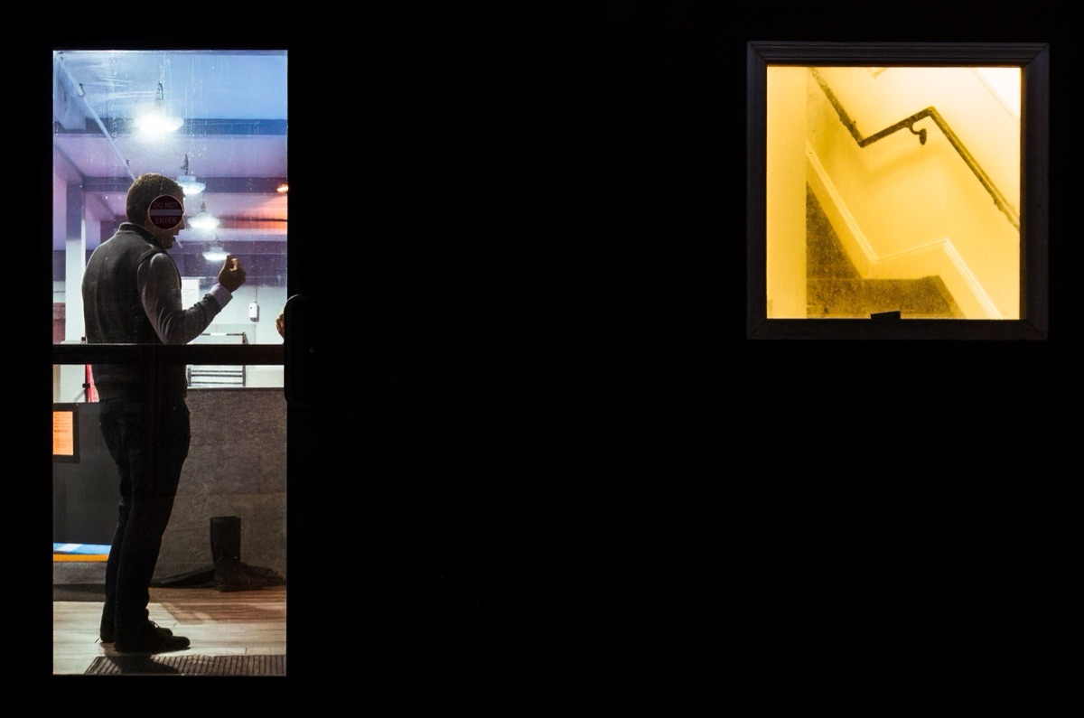 A masked figure framed in a lit doorway beside a glowing stairwell window at night