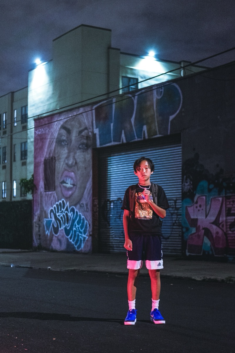 A young man standing before a floodlit graffiti mural on an empty street at night