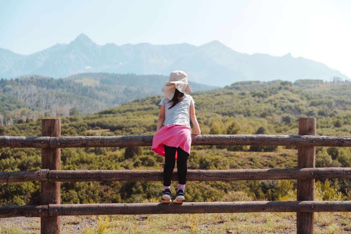 A girl in a sun hat climbing a split-rail fence to look out at the Rockies, Colorado