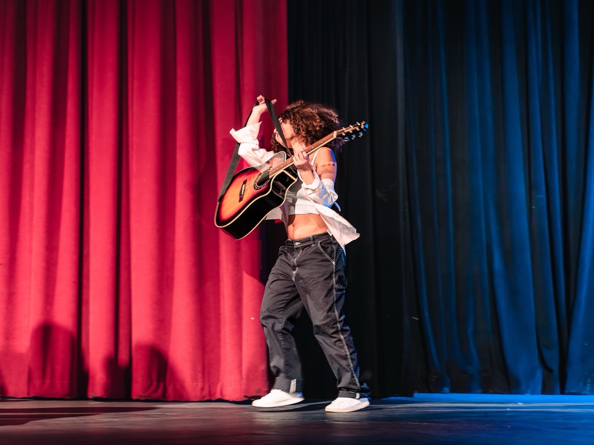 A student performing on acoustic guitar between red and blue stage curtains