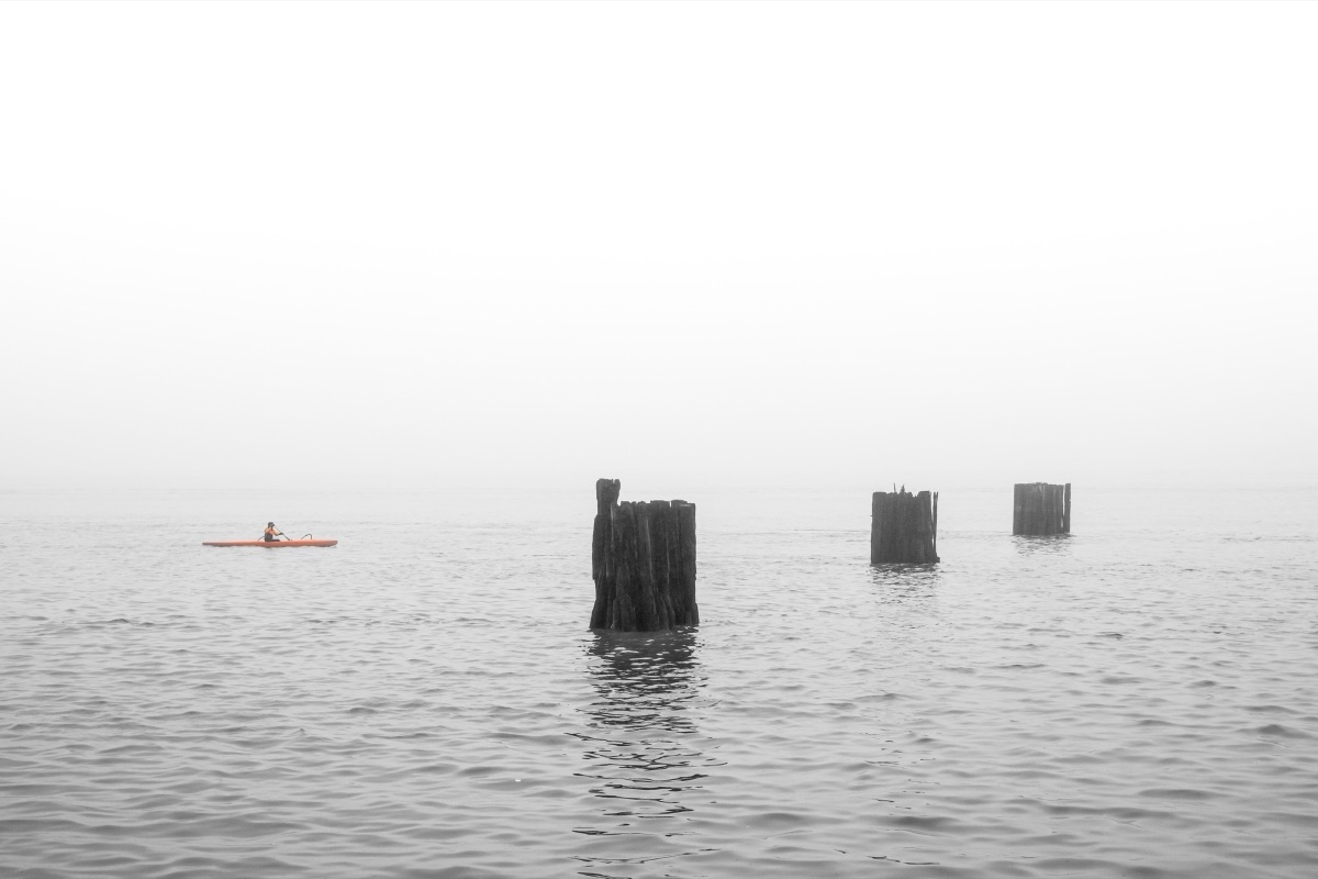 A lone kayaker paddling past old pier pilings in thick fog
