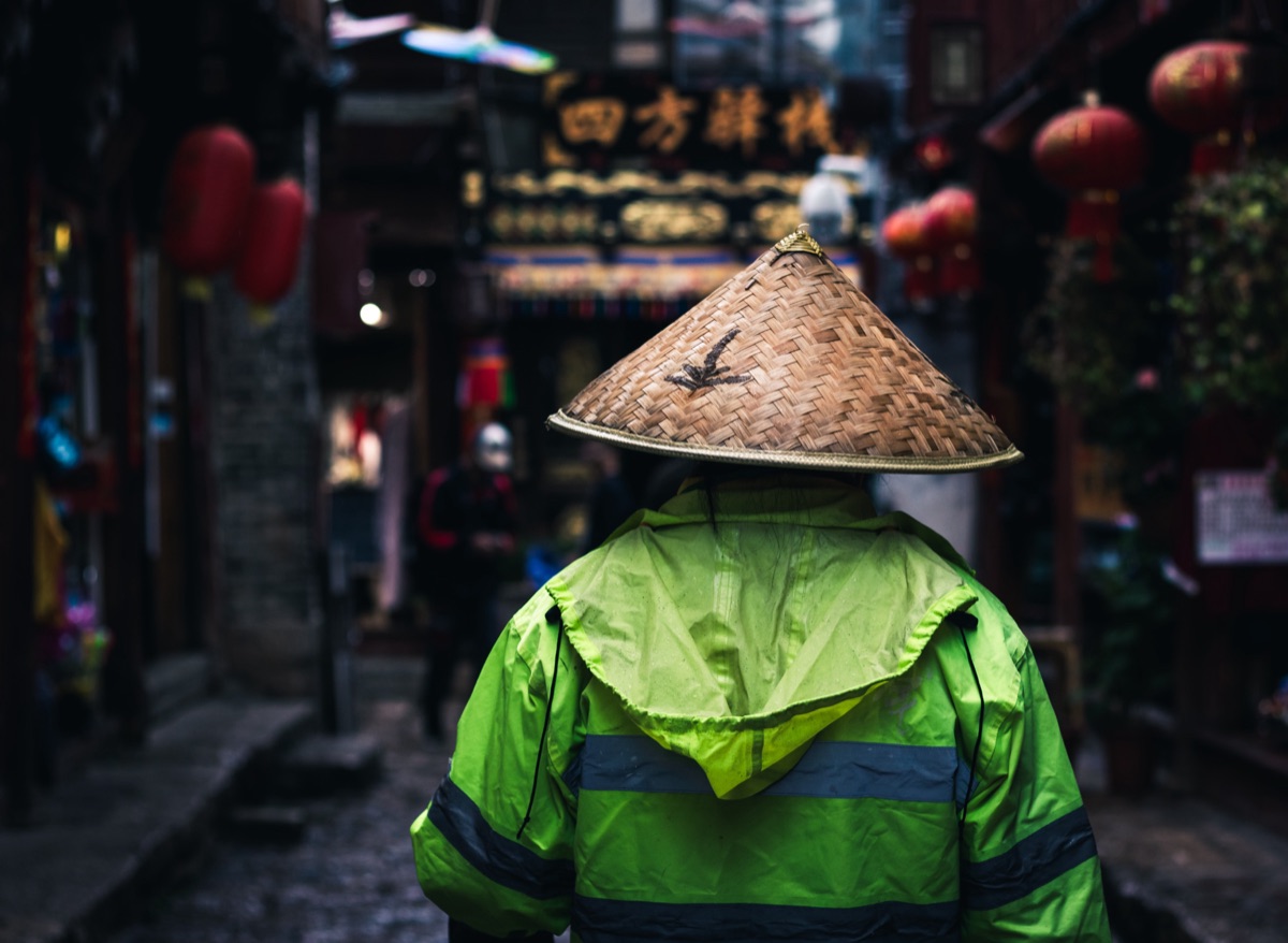 A street sweeper in a bamboo hat and high-visibility jacket walking a lantern-hung alley, China