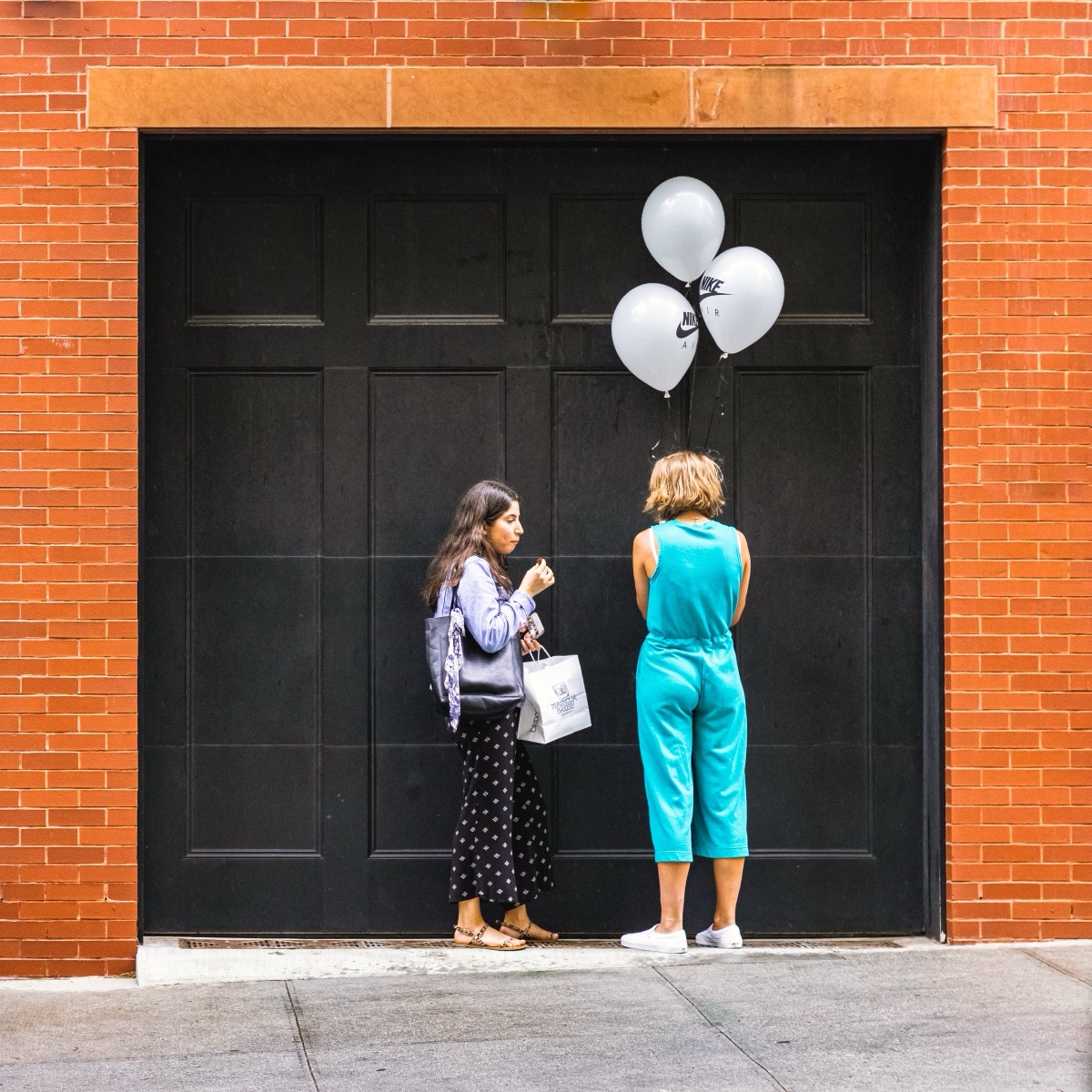 Two women talking with balloons outside a black loading door in a red brick wall