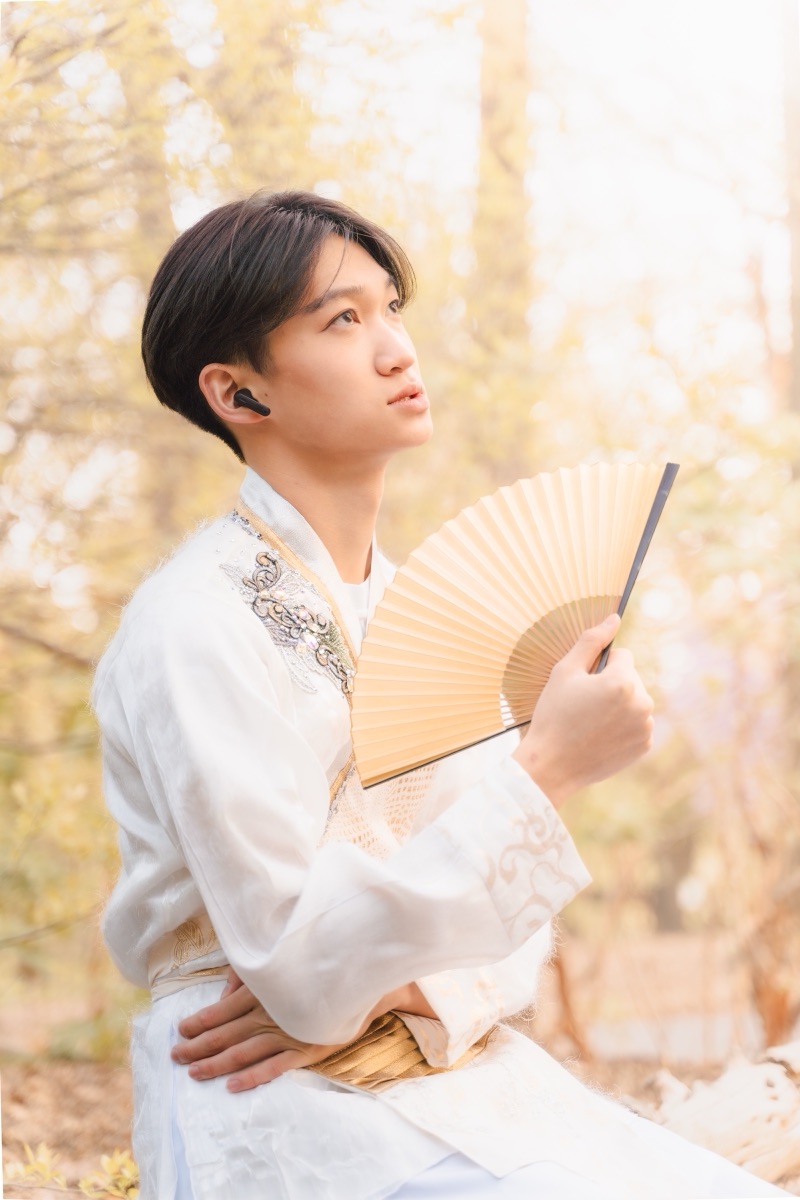 A young man in white hanfu holding a folding fan among sunlit ginkgo trees