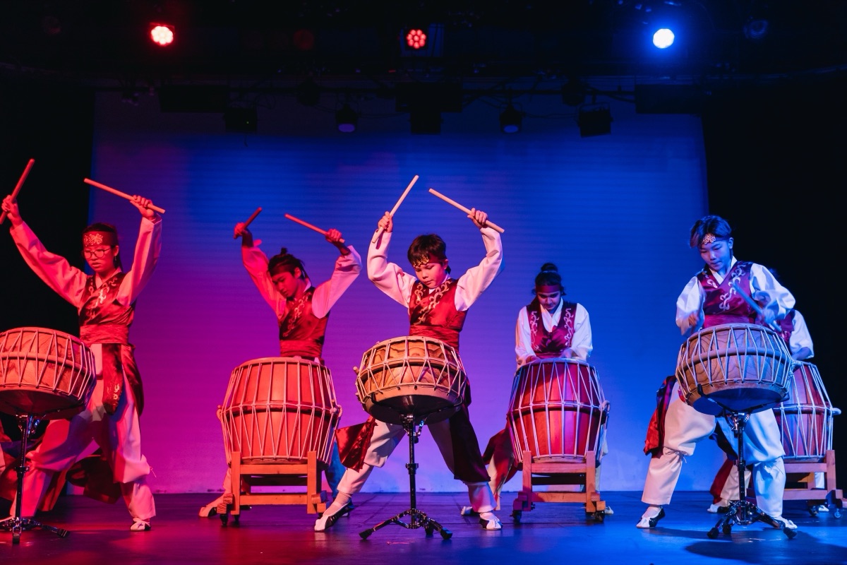 Korean drummers mid-strike on janggu and buk under magenta and blue stage light